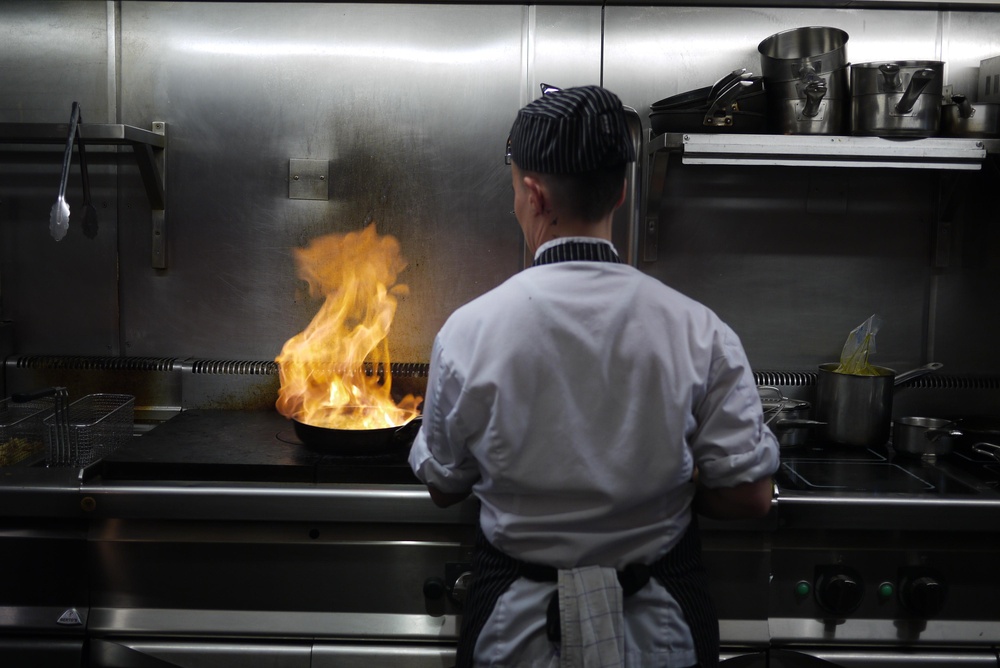 Back,Facing,Chef,In,A,Stainless,Steel,Restaurant,Kitchen,Cooking Chef cooking with open flames in a commercial kitchen setup.